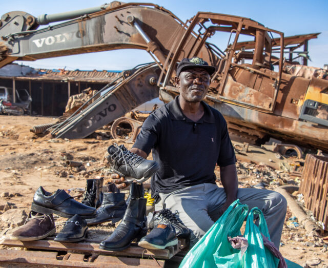 man sitting with a number of shoes with a rusty crane in the background