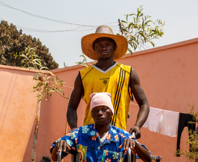 injured Didier sits in a wheelchair with na friend holding the wheelchair bars behind him