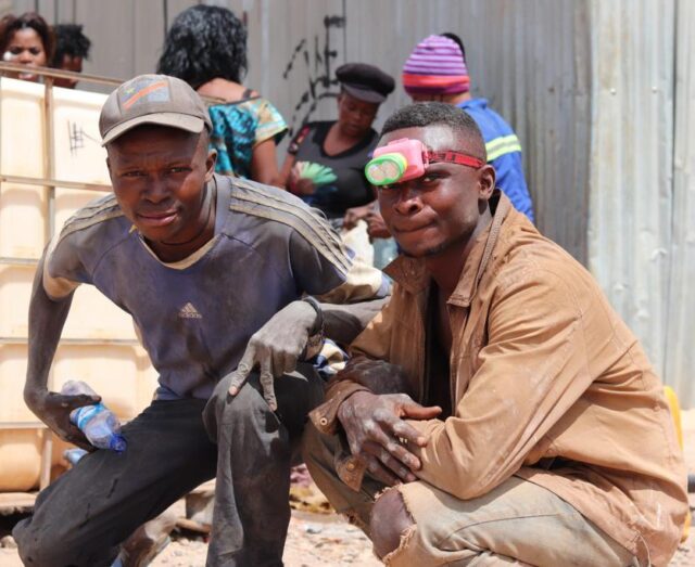 two black males posing for the camera, one wearing a head torch and covered in dust